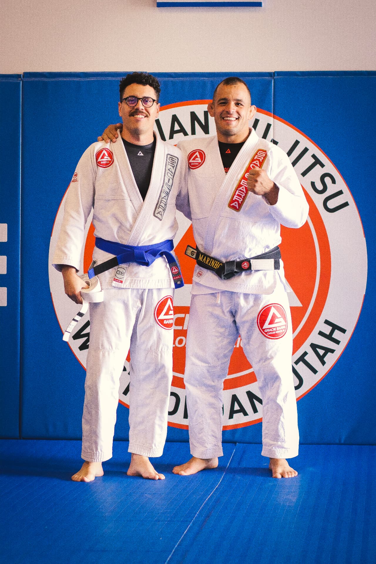 Two jiu jitsu practitioners in white gis smiling in front of a Gracie Barra Utah wall, one holding a new belt